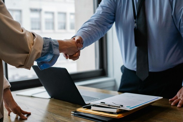 Person wearing a tie and blue dress shirt shaking hands with another person across a desk with a laptop and paperwork on it
